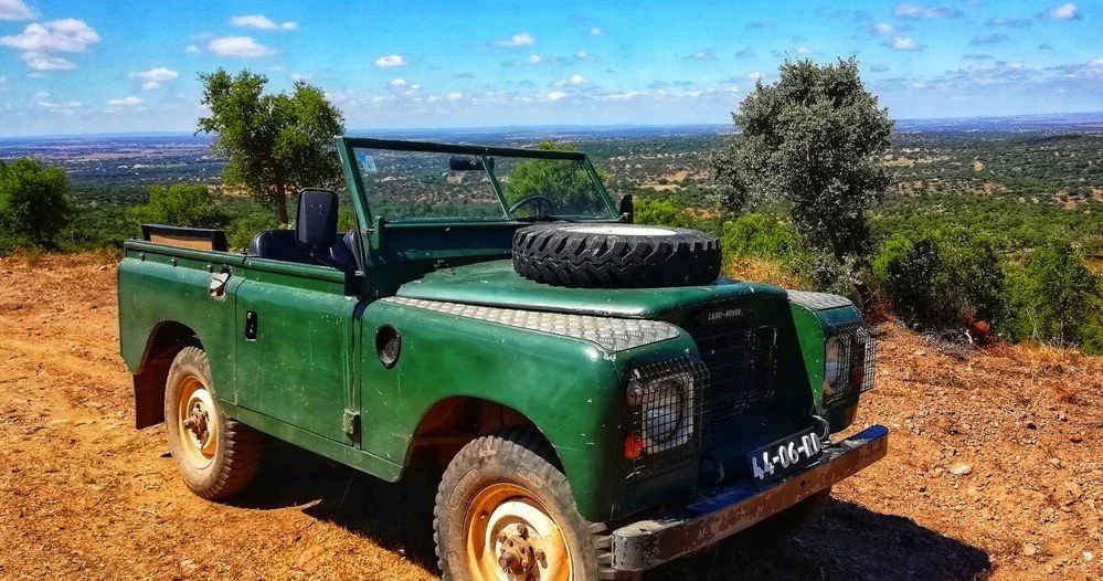 Land Rover clásico verde por una pista de tierra roja entre alcornoques