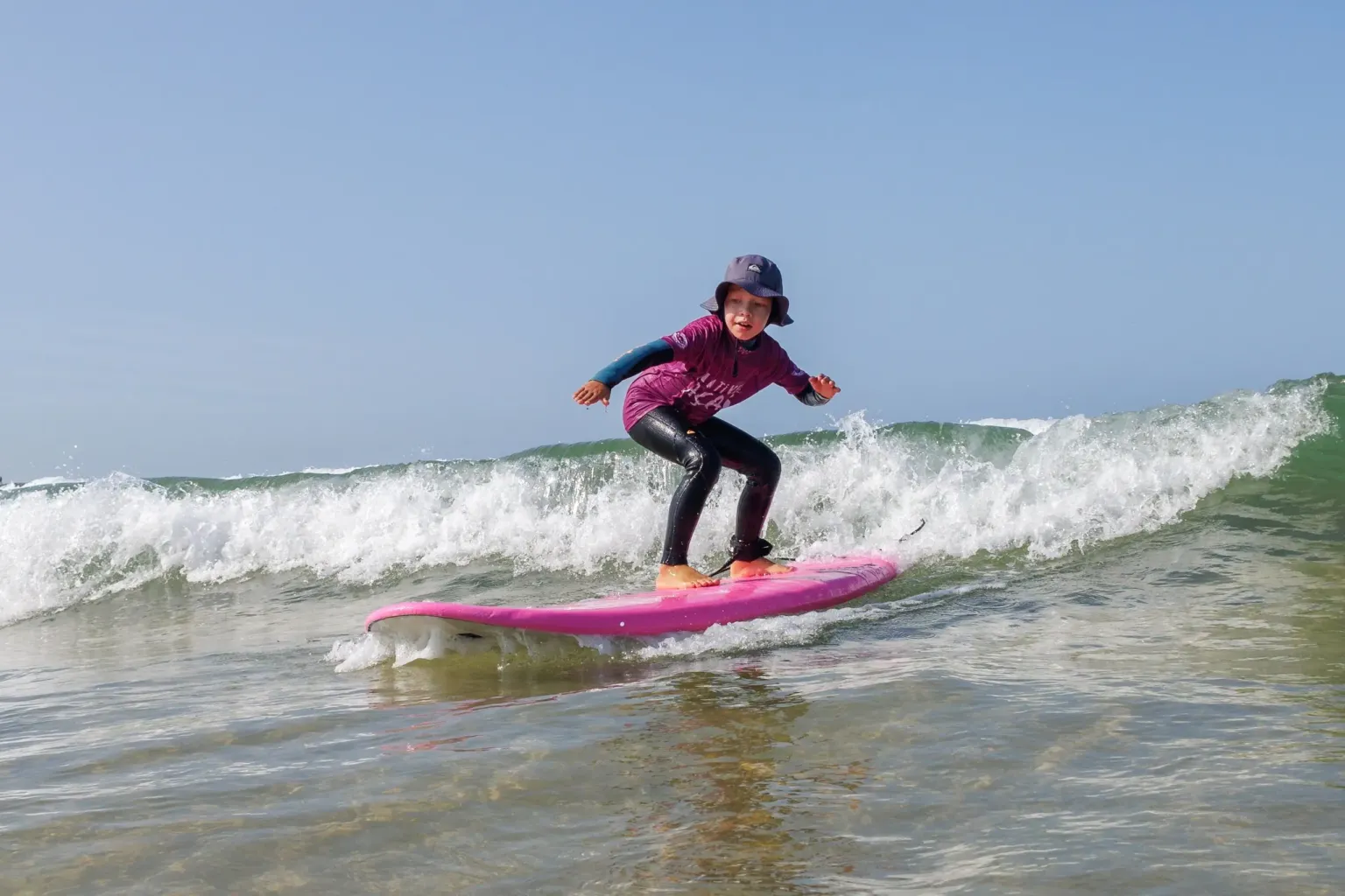 Child in a sun hat standing on a pink board on a wave