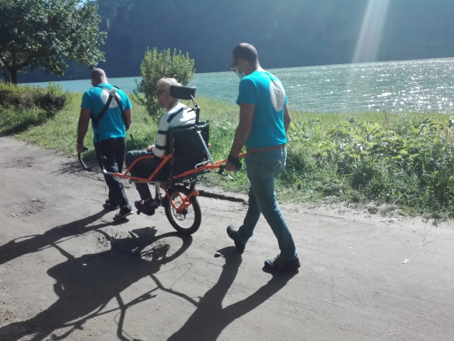 Two guides pushing a person seated in a one-wheeled trail chair along a lakeside road