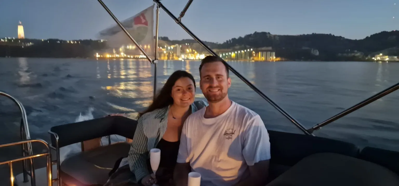 Couple smiling on a boat at night with town lights behind