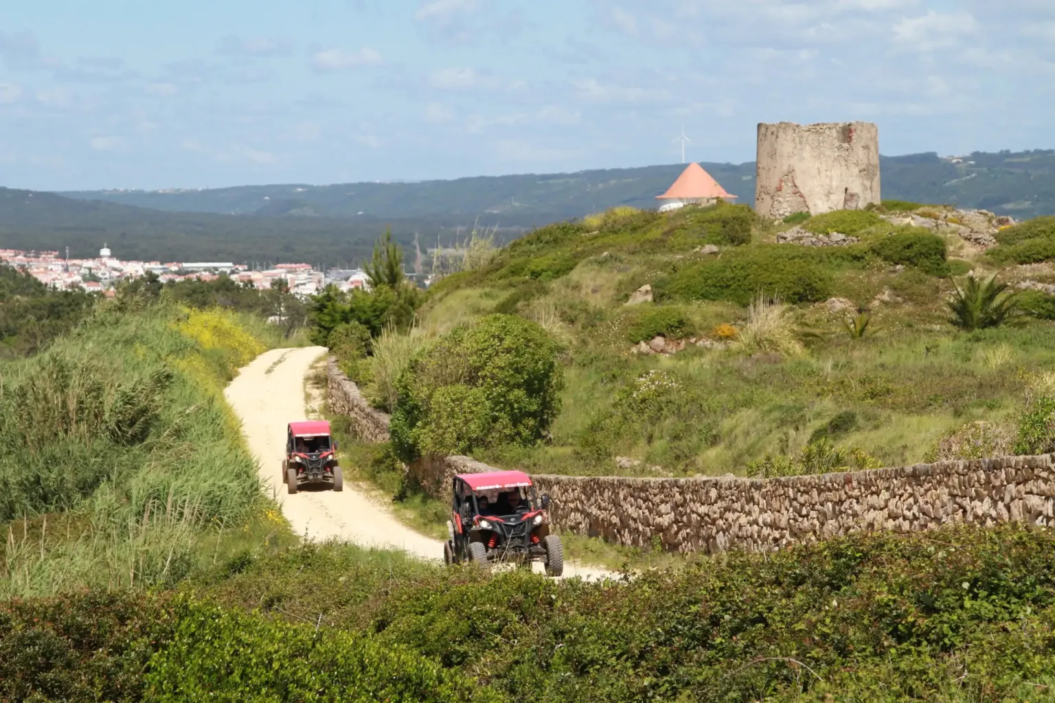 Two pink buggies on a dirt track winding through coastal scrub towards a hilltop tower