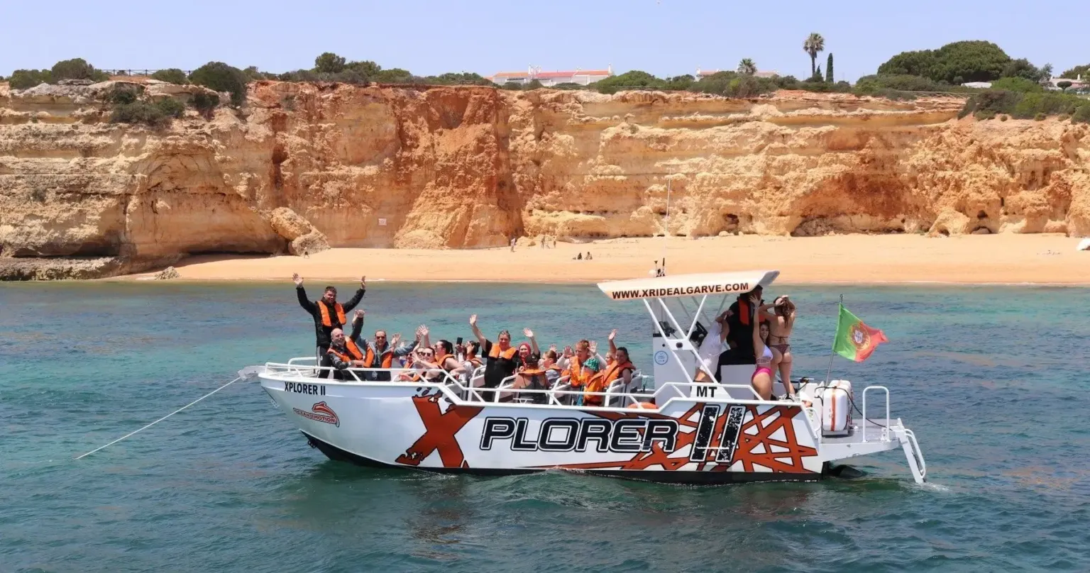 Passenger boat full of cheering guests anchored off a sandy beach below orange cliffs