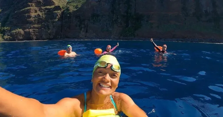 Smiling swimmer in a yellow cap with other swimmers behind, below sea cliffs