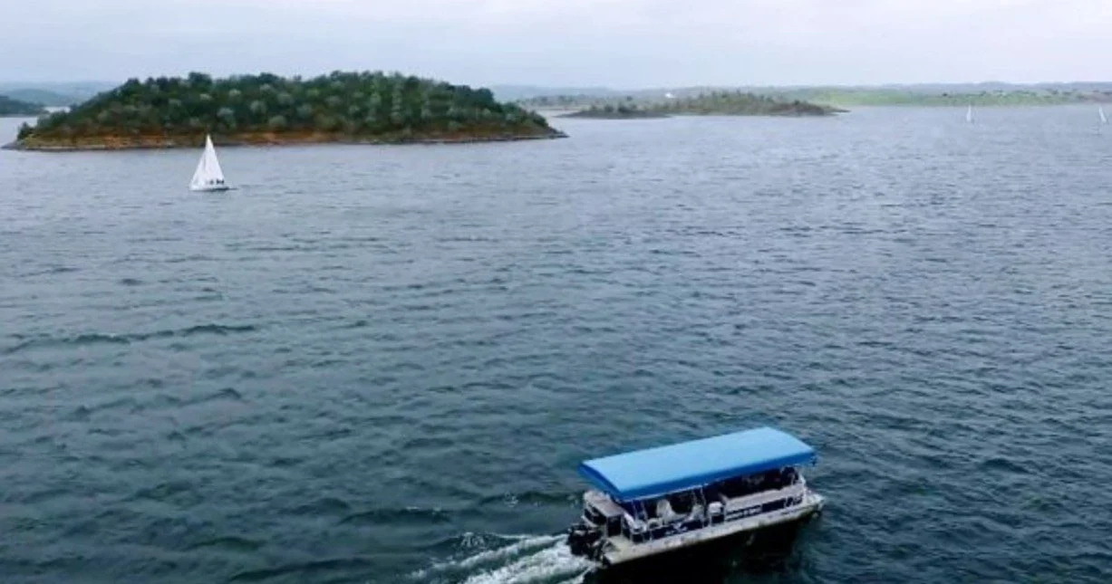 Small pontoon boat crossing a wide lake with a wooded island and a sailboat