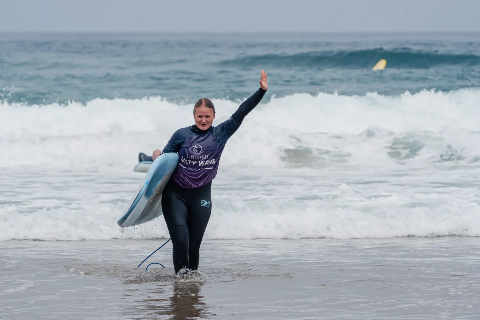 Surfer in a wetsuit raising an arm while carrying a board out of the sea