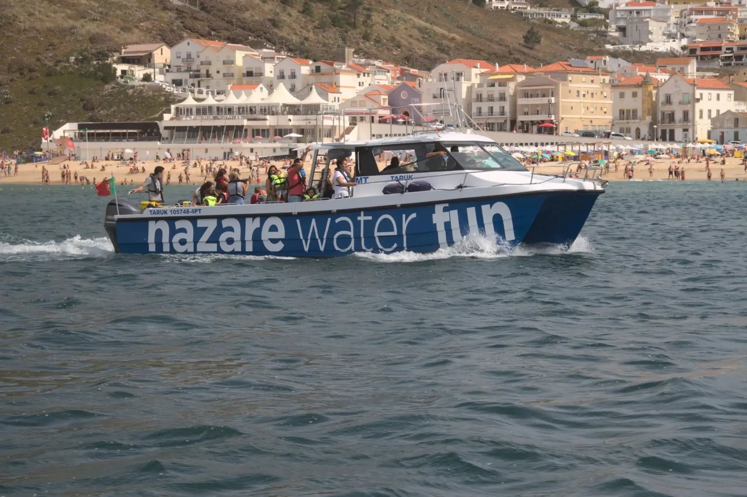 Boat named Nazaré Water Fun cruising off a crowded beach and town