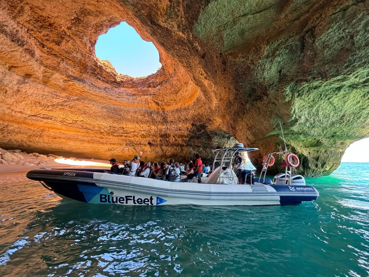 Barco insuflável com passageiros a entrar numa gruta marítima de arenito com teto aberto