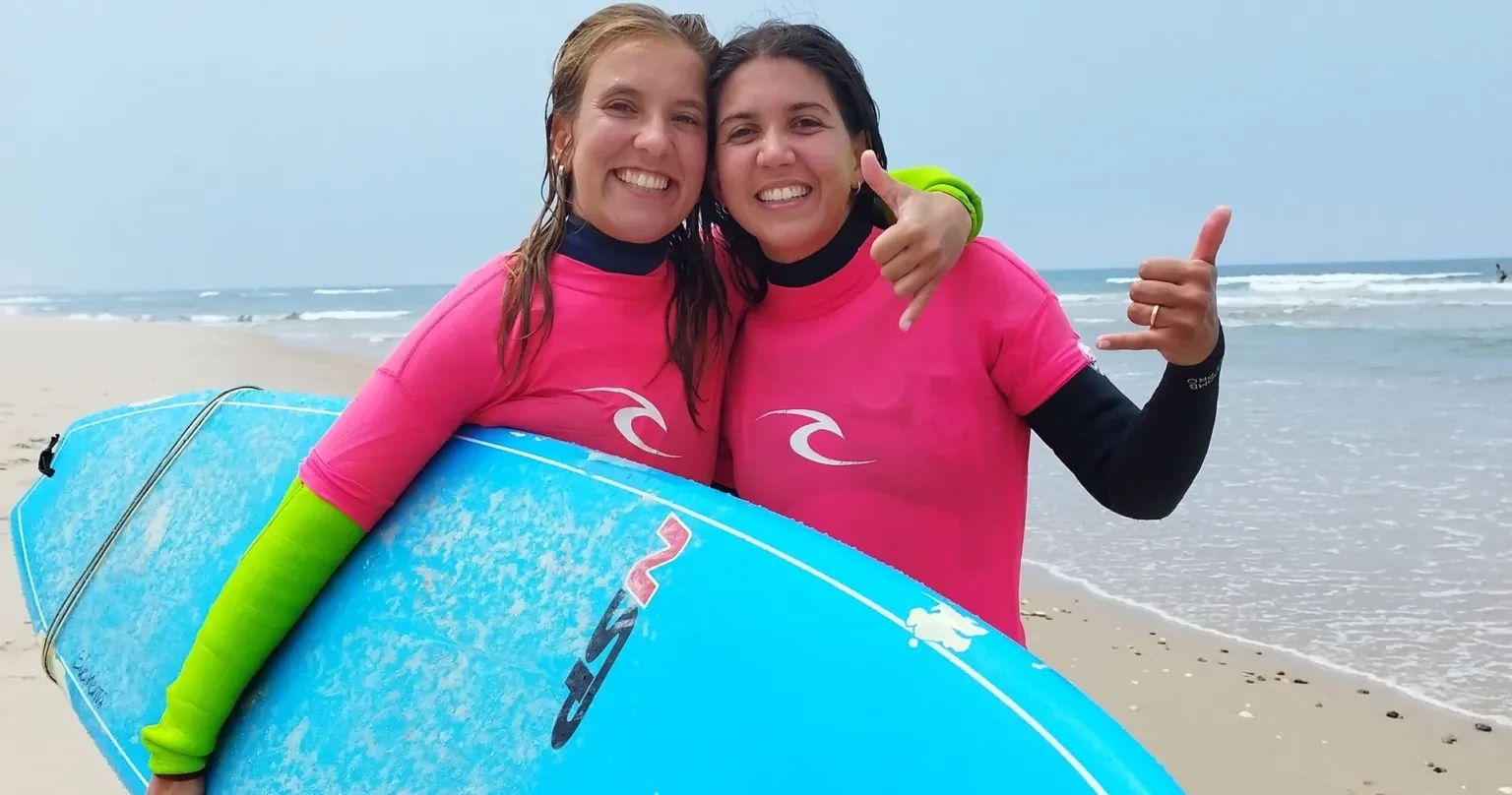 Two smiling surfers in pink rash vests holding a blue board on the beach