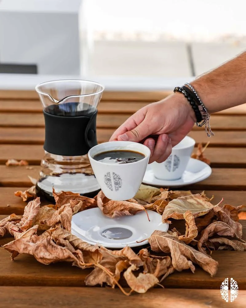 Hand placing a coffee cup beside dried leaves on a wooden table