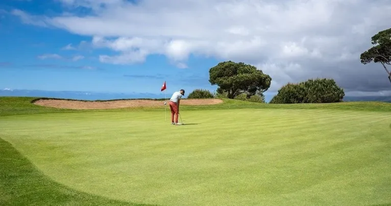 Golfer putting on a green beside a sand bunker under cloudy sky