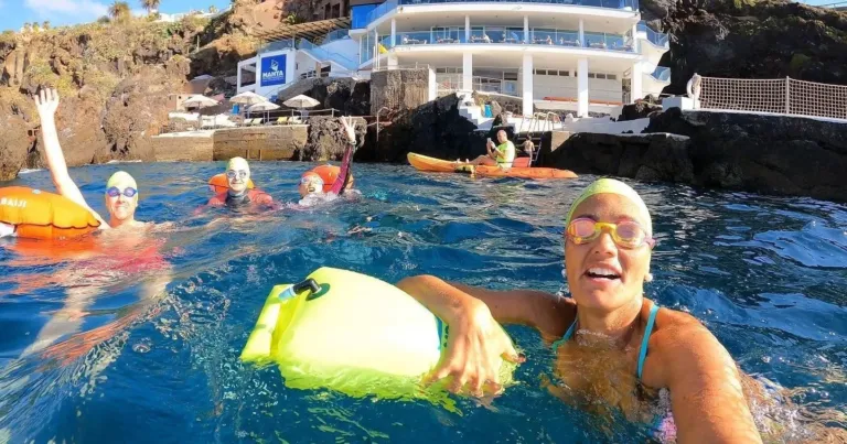 Smiling swimmer holding a yellow tow float in the sea, with a seaside hotel behind