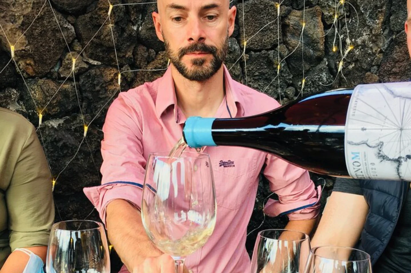 Man in a pink shirt pouring wine at a tasting among lava stone walls