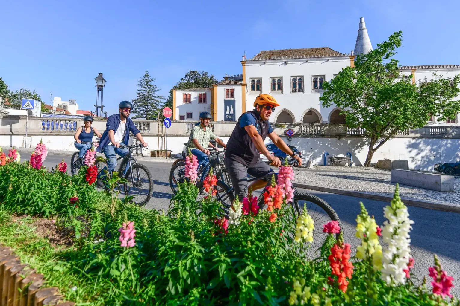 Cyclists riding past flower beds towards a palace with a conical chimney