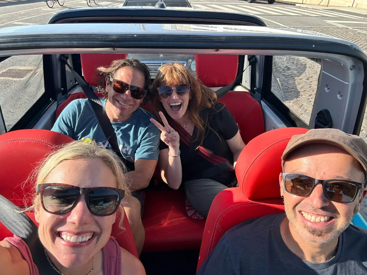 Group of four smiling travellers in sunglasses taking a selfie in a red-seated open vehicle