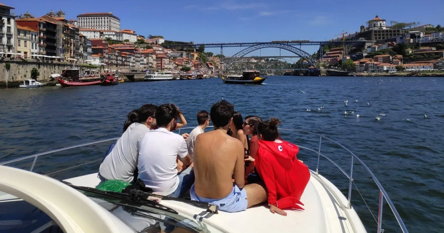 Passengers on a yacht's bow looking towards a riverside city and a bridge