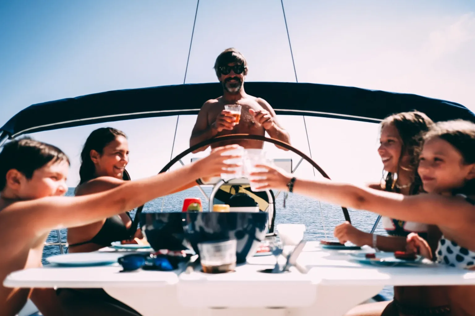 Family toasting around a table on a sailboat while a man steers