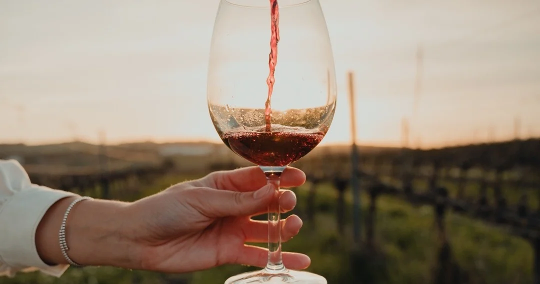 Hand holding a glass as red wine is poured, with vineyards at sunset