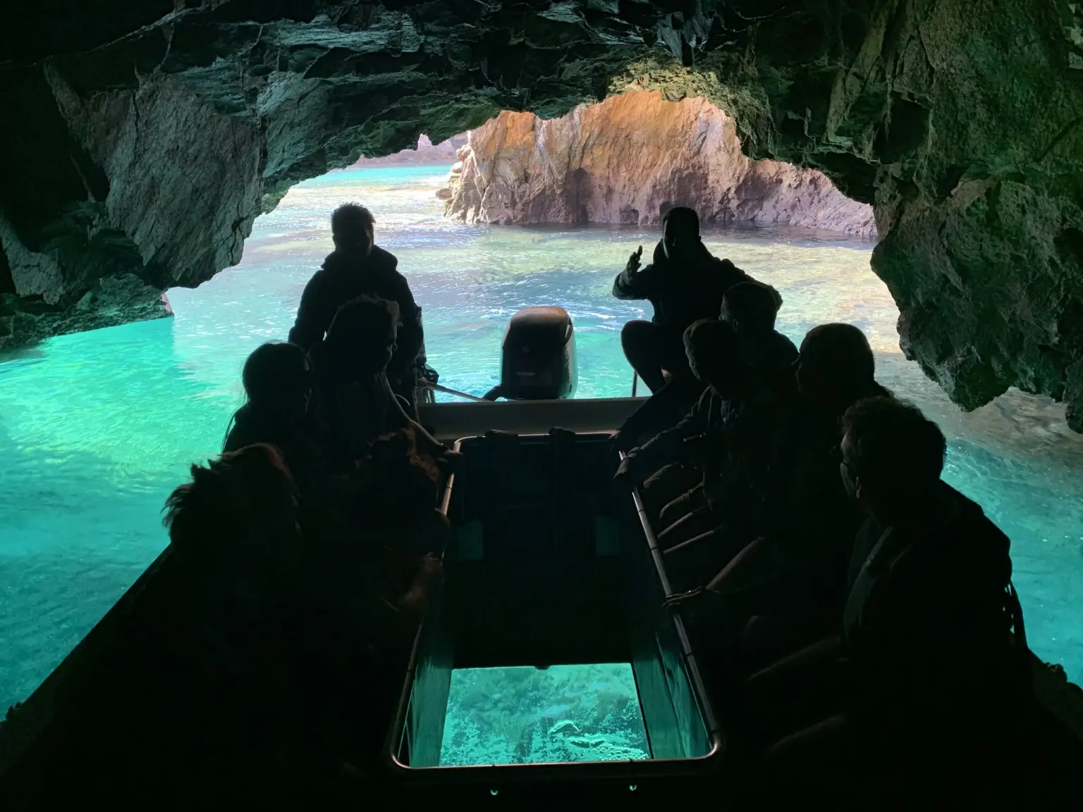 Silhouetted passengers in a glass-bottom boat entering a green-lit sea cave