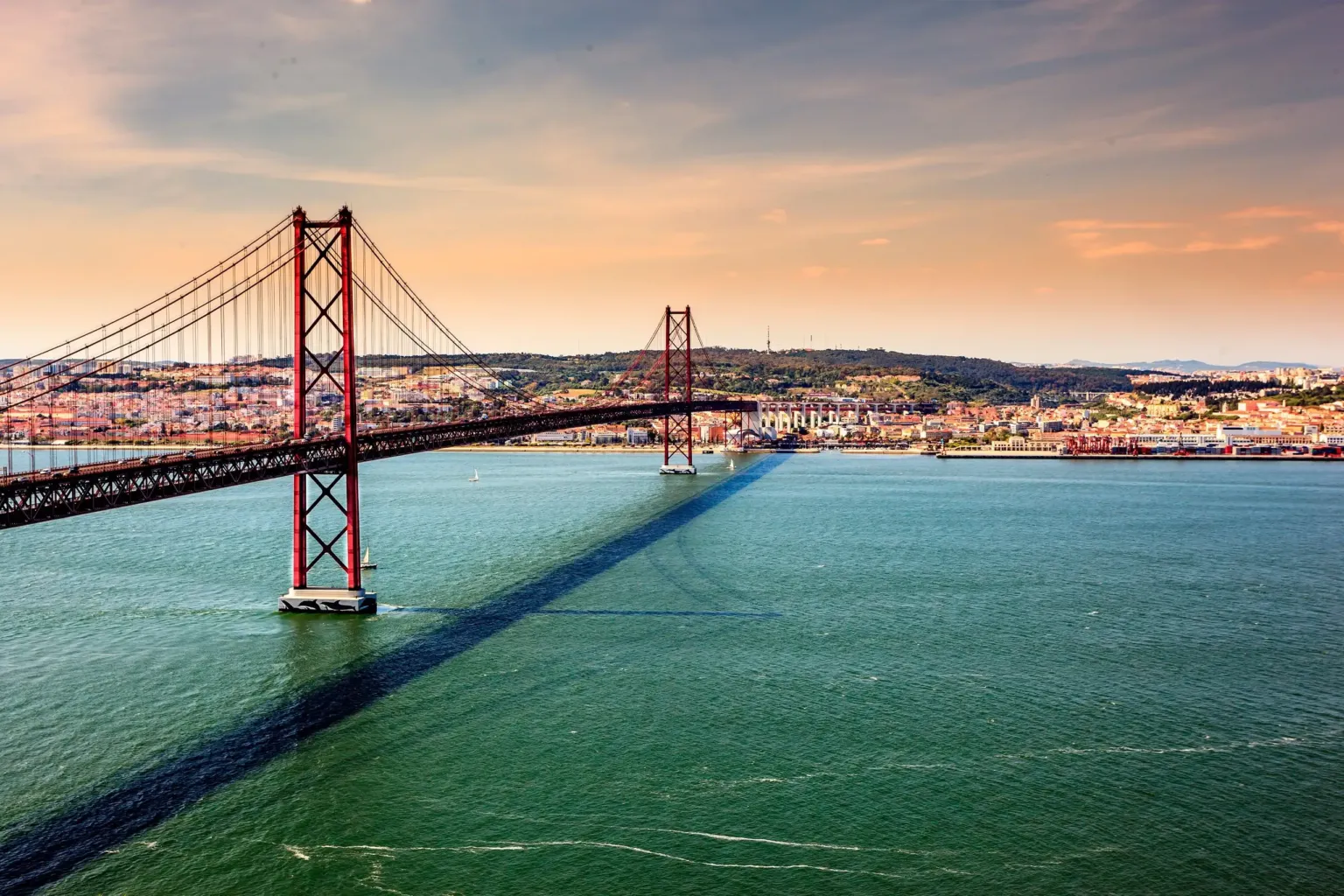Red suspension bridge spanning green water with the city in the distance