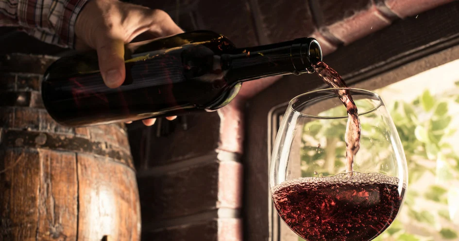 Red wine being poured into a glass beside wooden barrels