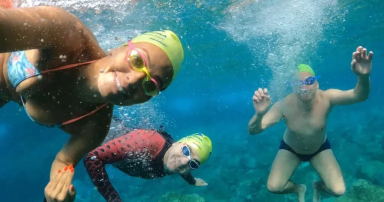 Swimmers in caps and goggles smiling underwater over rocks