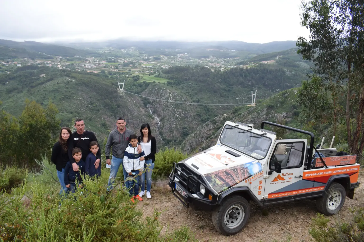 Family beside an open-top jeep with a view over a valley and suspension bridge