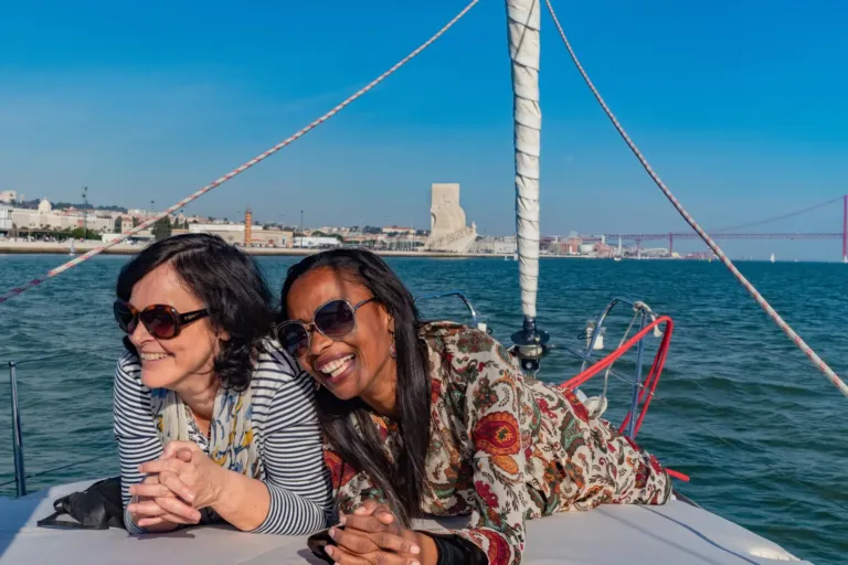 Two women laughing on the foredeck of a sailboat, with a monument and bridge behind