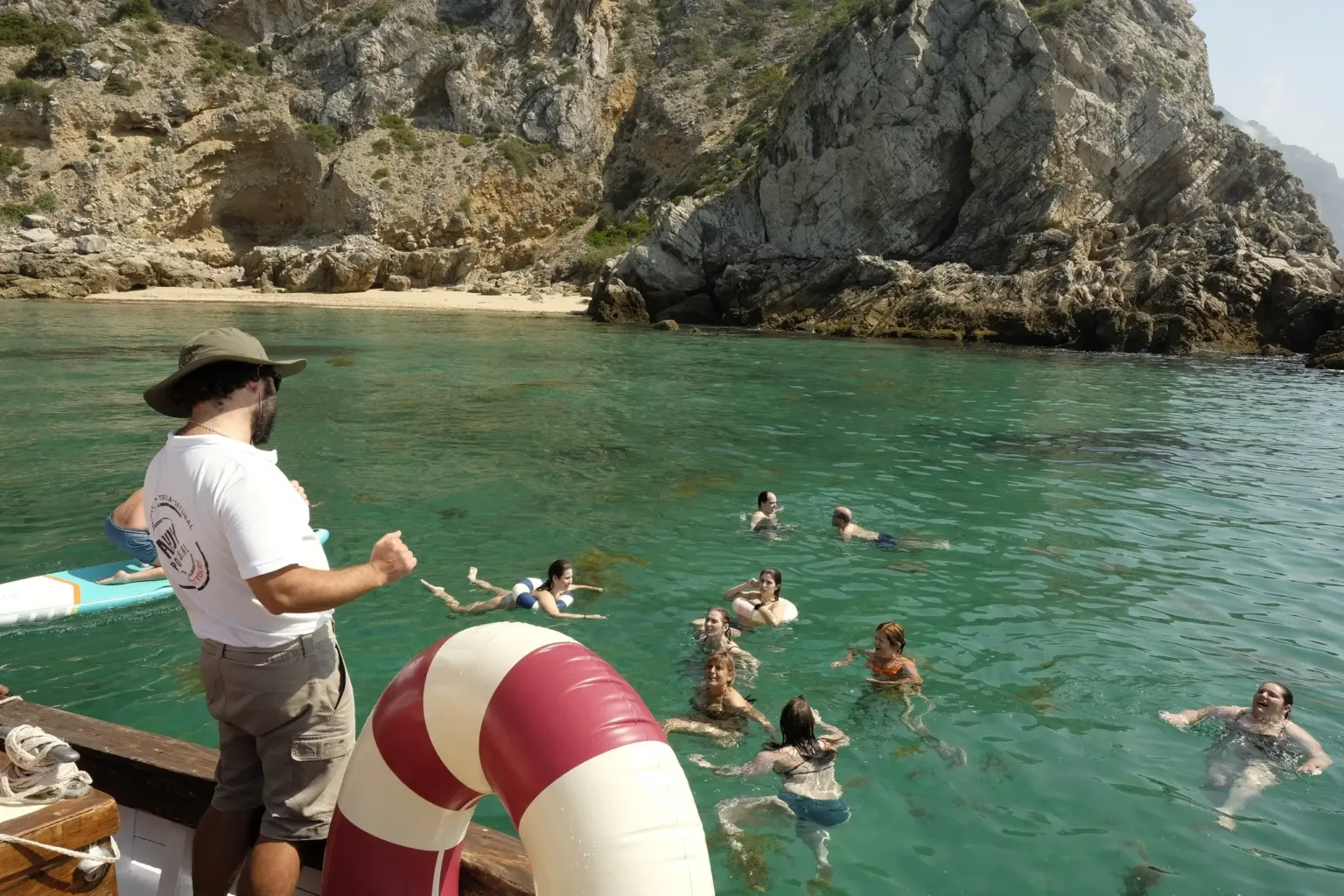 Skipper looking on as guests swim in clear green water beside a cliff