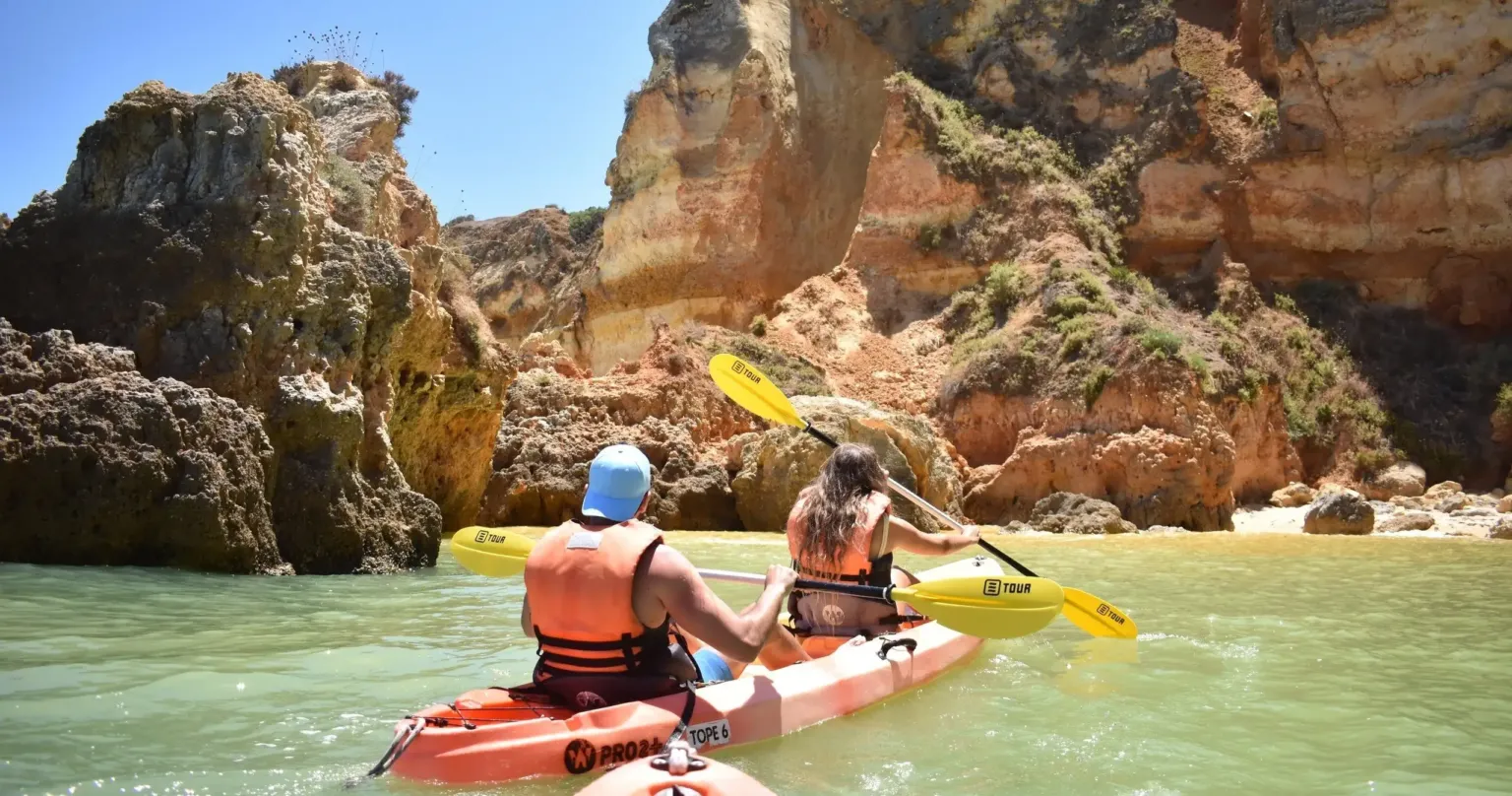 Two kayakers paddling past golden cliffs in clear green water