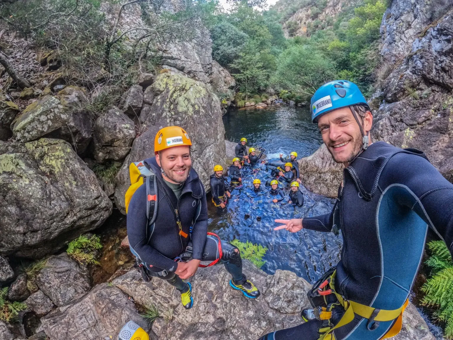 Two canyoners in helmets smiling, with a group in a river gorge behind