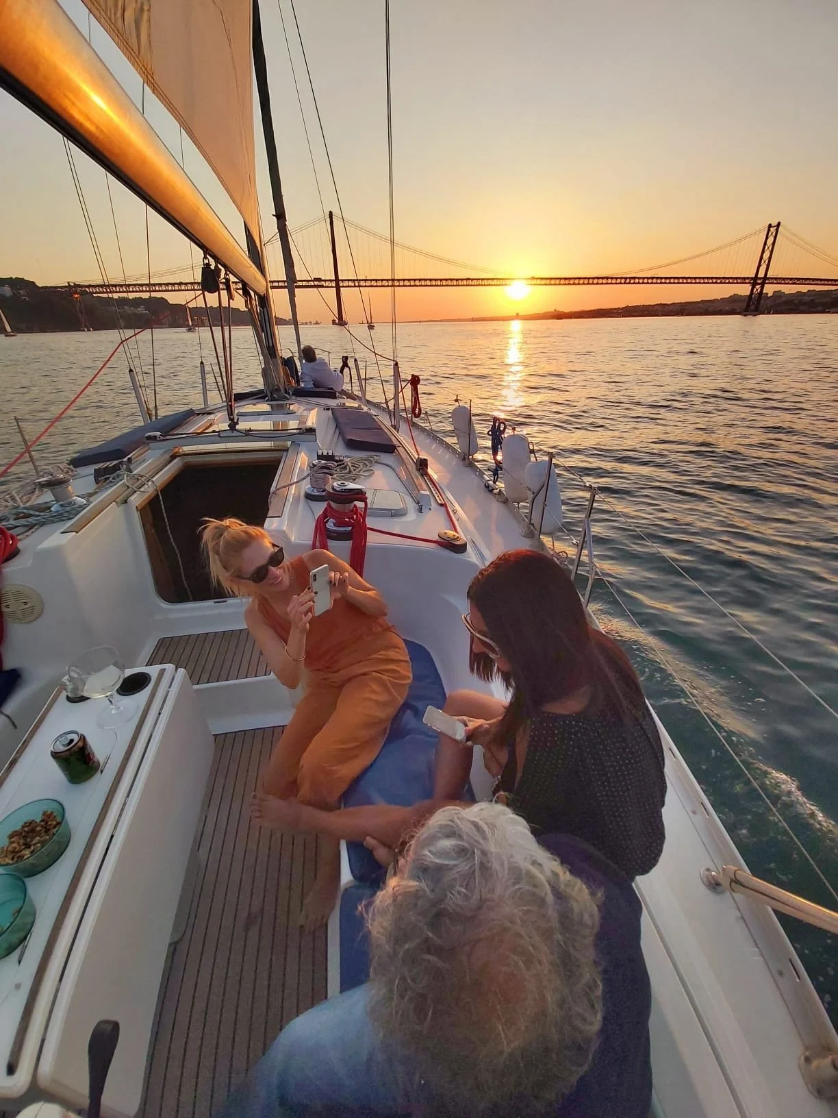 Guests relaxing on the deck of a sailboat at sunset with a bridge ahead
