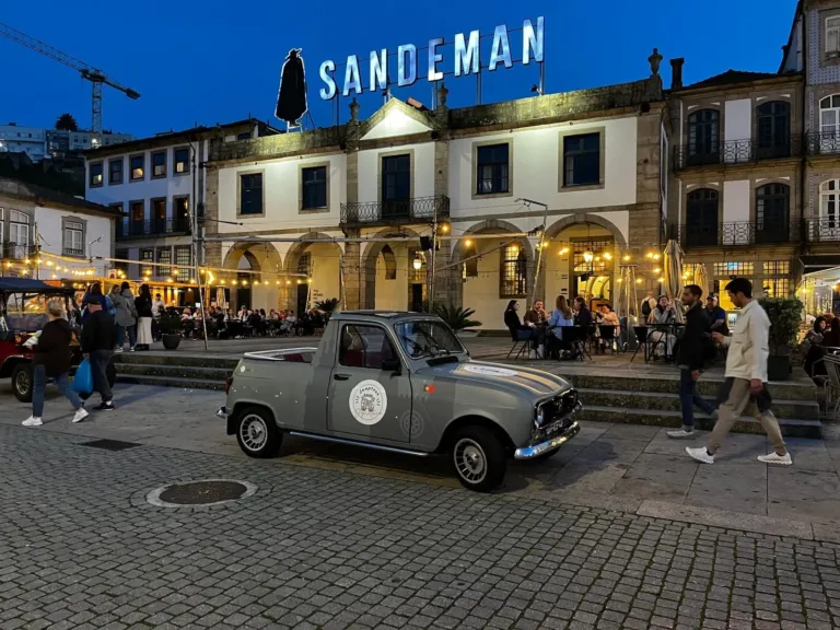 Small vintage Renault parked on a city square at dusk, below a lit 'Sandeman' sign