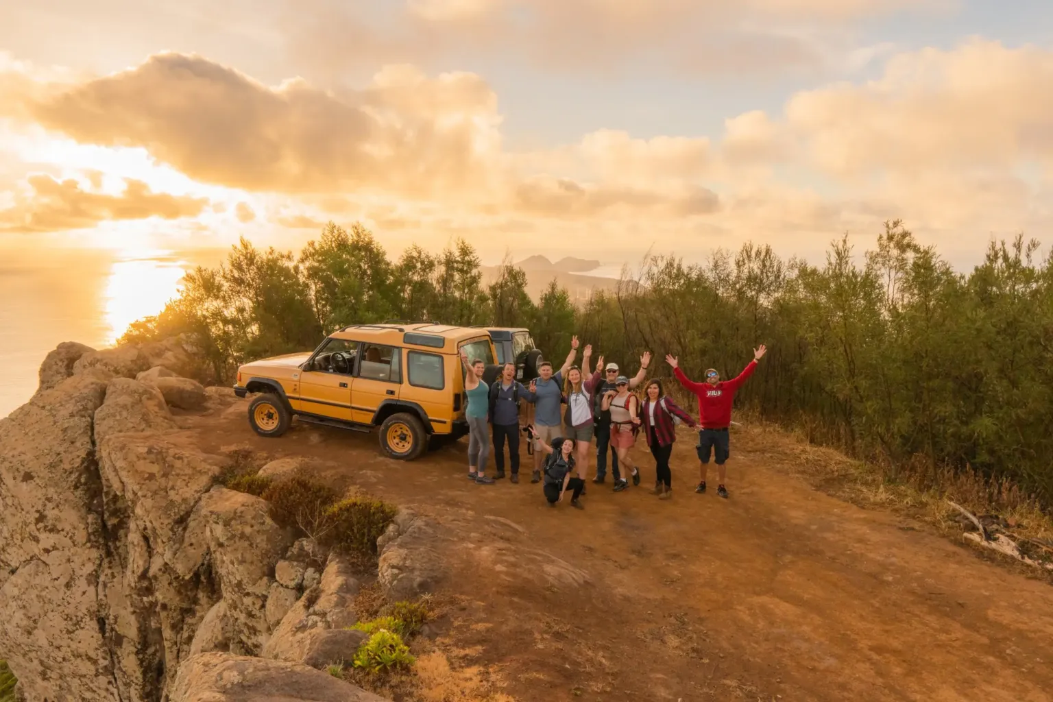 Group with raised arms beside a yellow 4x4 on a cliff edge at sunset